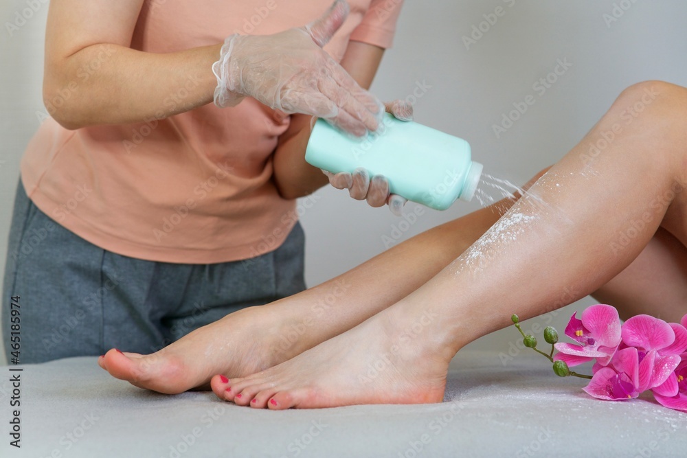 Fototapeta premium Cosmetologist woman is sprinkling talcum powder on young girl's leg before the epilation or depilation procedure. Girl is lying on a couch in a beauty salon on procedure shugaring with orchid flower