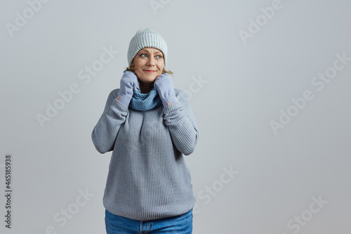 mature woman in a gray sweater and knitted hat wrapped herself in a scarf from the cold. Isolated on gray background © evafesenuk