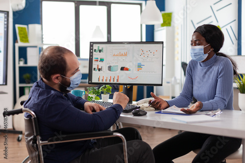 Paralyzed businessman in wheelchair wearing protective face mask to prevent infection with coronavirus working in startup business office. Multi-ethnic workteam discussing company strategy