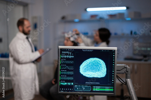 Monitor with brain activity on screen in neurology medical laboratory. In background team of neurologist adjusting eeg headset of woman patient monitoring nervous system during neuroscience experiment photo