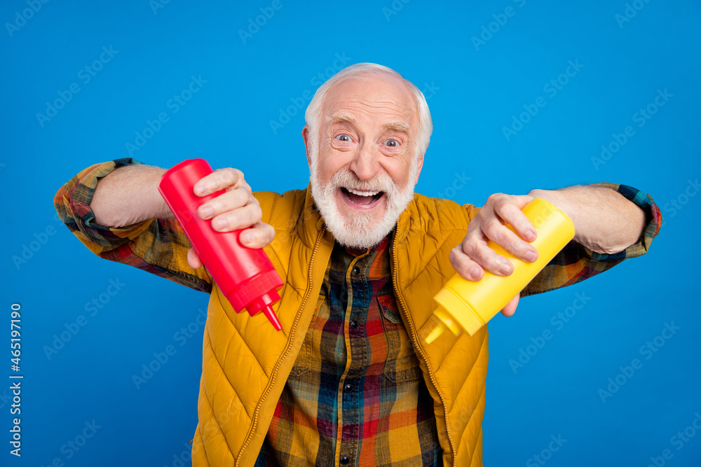 Portrait of attractive cheerful grey-haired man holding bottles ketchup adding dish meal isolated over bright blue color background