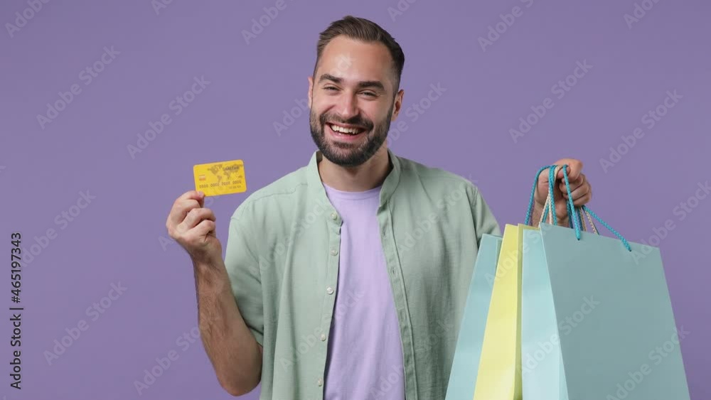 Smiling satisfied happy young bearded man 20s years old wears mint shirt holding package bags with purchases after shopping credit bank card isolated on plain light purple background studio portrait