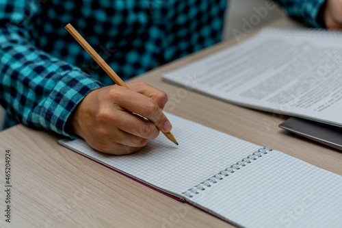 A woman in a plaid shirt writes with a pencil in a notebook and looks into a paper report. The right female hand is in focus. Horizontal view.