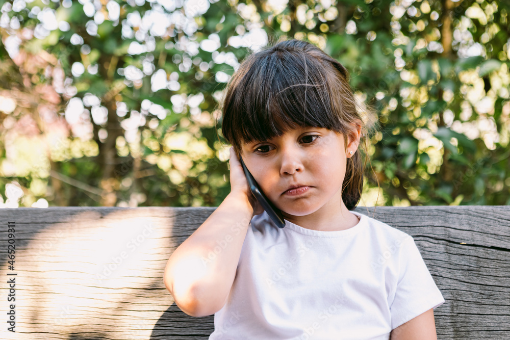 Small girl with black hair, wearing a white T-shirt, sitting on a bench in a park, talking on the mobile phone, with a melancholic gesture. Small girl with black hair, wearing a white T-shirt, sitting on a bench in a park, talking on the mobile phone, with a melancholic gesture.