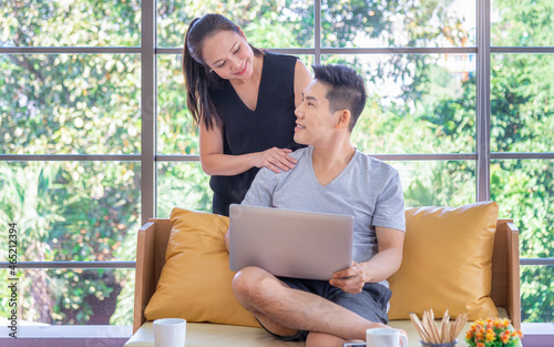 Beautiful young asian couple using laptop on the sofa at home. Young man working from home on laptop. Woman is smiling at her. Happy couple enjoying work from home  © sutthinon602