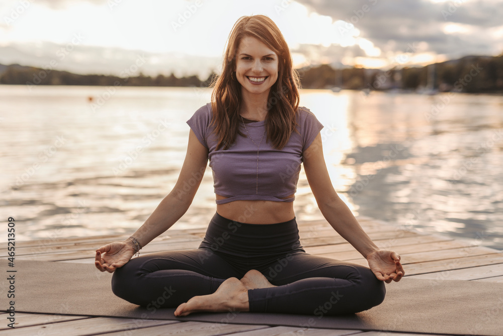 Smiling woman practicing lotus position while sitting on exercise mat by lake during sunset