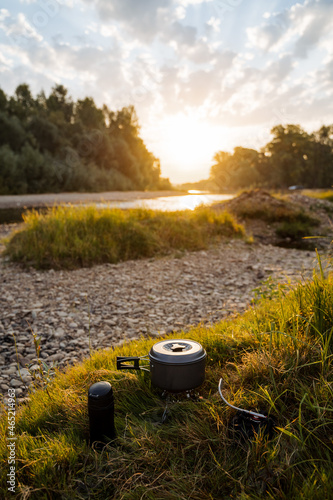 Sunset at the rocky bank of the river in summer. Meal tourist in nature, cooking in the wild. Iron pot and thermos in the grass. Hiking and active rest.