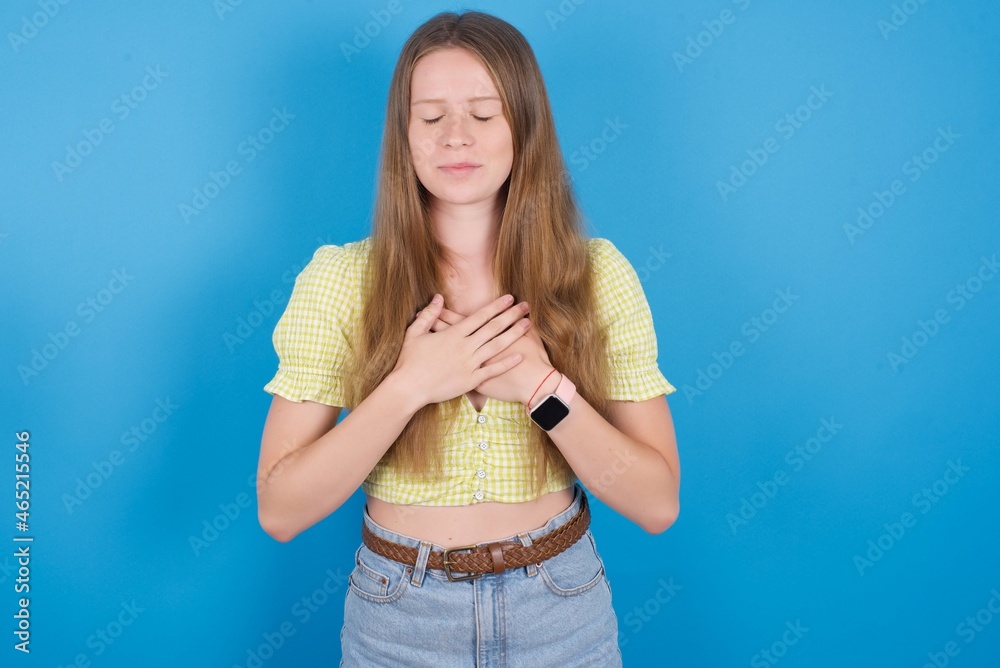 young ukranian girl wearing yellow t-shirt over blue backaground closes eyes and keeps hands on chest near heart, expresses sincere emotions, being kind hearted and honest. young ukranian girl wearing yellow t-shirt over blue backaground closes eyes and keeps hands on chest near heart, expresses sincere emotions, being kind hearted and honest.