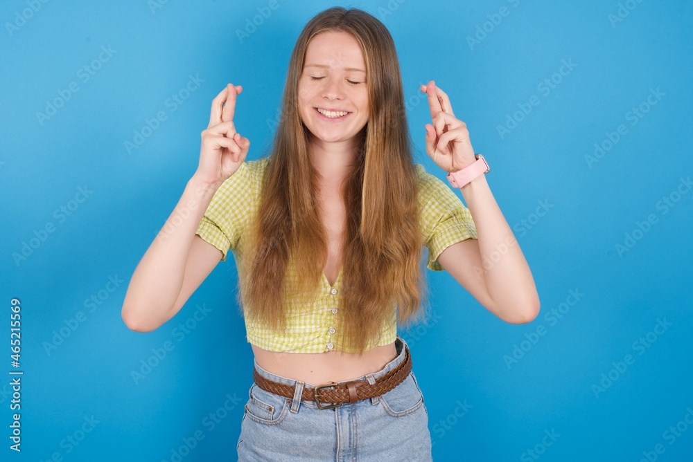 Fototapeta premium Joyful young ukranian girl wearing yellow t-shirt over blue backaground clenches teeth, raises fingers crossed, makes desirable wish, waits for good news, I have to win.