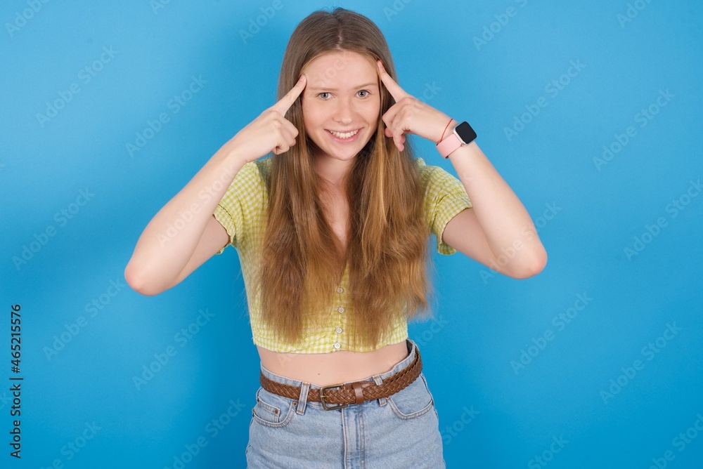 young ukranian girl wearing yellow t-shirt over blue backaground concentrating hard on an idea with a serious look, thinking with both index fingers pointing to forehead.