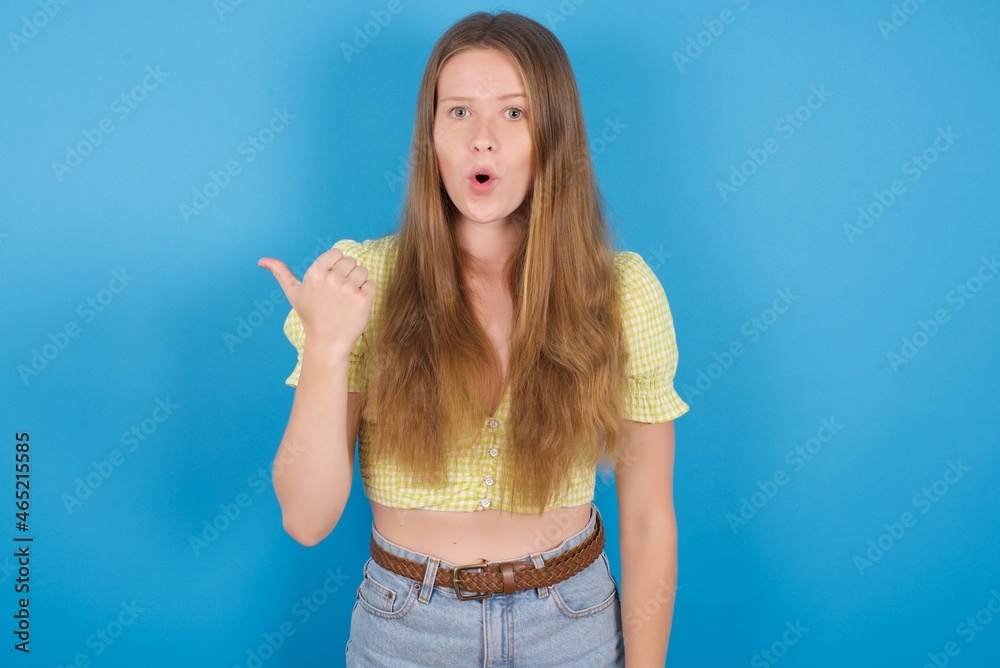 Shocked young ukranian girl wearing yellow t-shirt over blue backaground points with thumb away, indicates something. Check this out. Advertisement concept. Shocked young ukranian girl wearing yellow t-shirt over blue backaground points with thumb away, indicates something. Check this out. Advertisement concept.