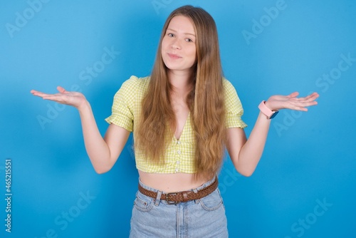 Maybe yes or no. Doubtful young ukranian girl wearing yellow t-shirt over blue backaground shrugs shoulders in bewilderment, tries to make decision puzzled what he wants. Hesitation and uncertainty. photo