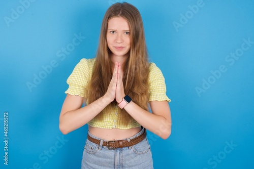 young ukranian girl wearing yellow t-shirt over blue backaground keeps palms pressed together in front of her having regretful look, asking for forgiveness. Forgive me please. photo