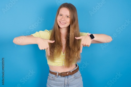 young ukranian girl wearing yellow t-shirt over blue backaground with positive expression, points down with both index fingers, has broad interested smile. Look there, please.