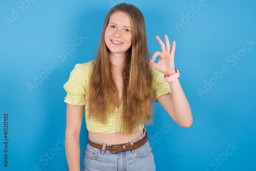 Glad attractive young ukranian girl wearing yellow t-shirt over blue backaground shows ok sign with hand as expresses approval, has cheerful expression, being optimistic.