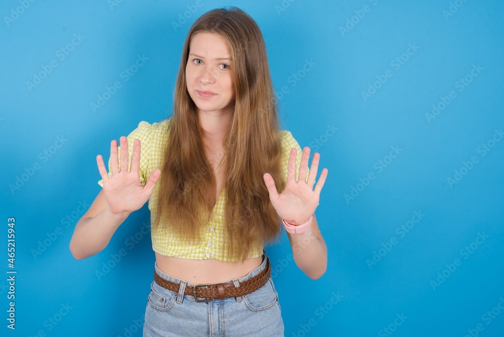 young ukranian girl wearing yellow t-shirt over blue backaground doing stop sing with palm of the hand. Warning expression with negative and serious gesture on the face. young ukranian girl wearing yellow t-shirt over blue backaground doing stop sing with palm of the hand. Warning expression with negative and serious gesture on the face.