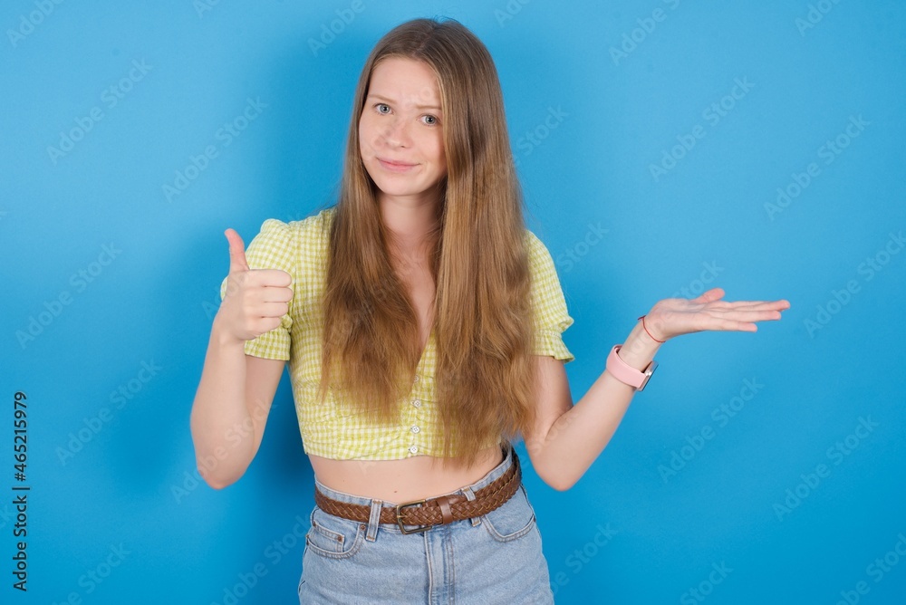 Happy cheerful young ukranian girl wearing yellow t-shirt over blue backaground showing thumb up and pointing with the other hand. I recommend this. Happy cheerful young ukranian girl wearing yellow t-shirt over blue backaground showing thumb up and pointing with the other hand. I recommend this.