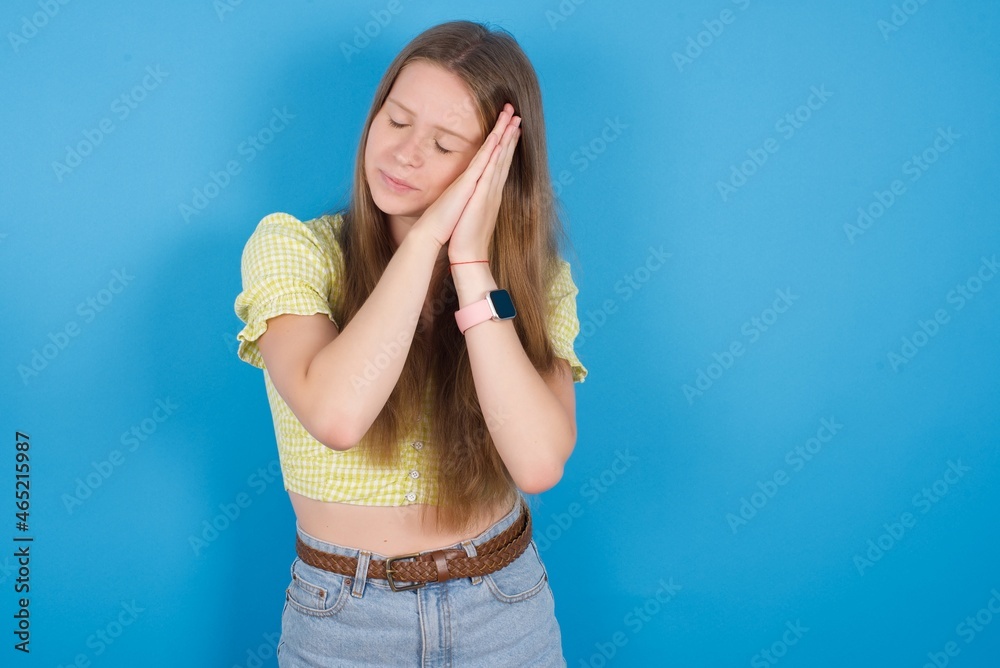 Relax and sleep time. Tired young ukranian girl wearing yellow t-shirt over blue backaground with closed eyes leaning on palms making sleeping gesture.