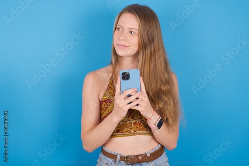 Portrait of young ukranian girl wearing tank top over blue backaground with dreamy look, thinking while holding smartphone. Tries to write up a message.