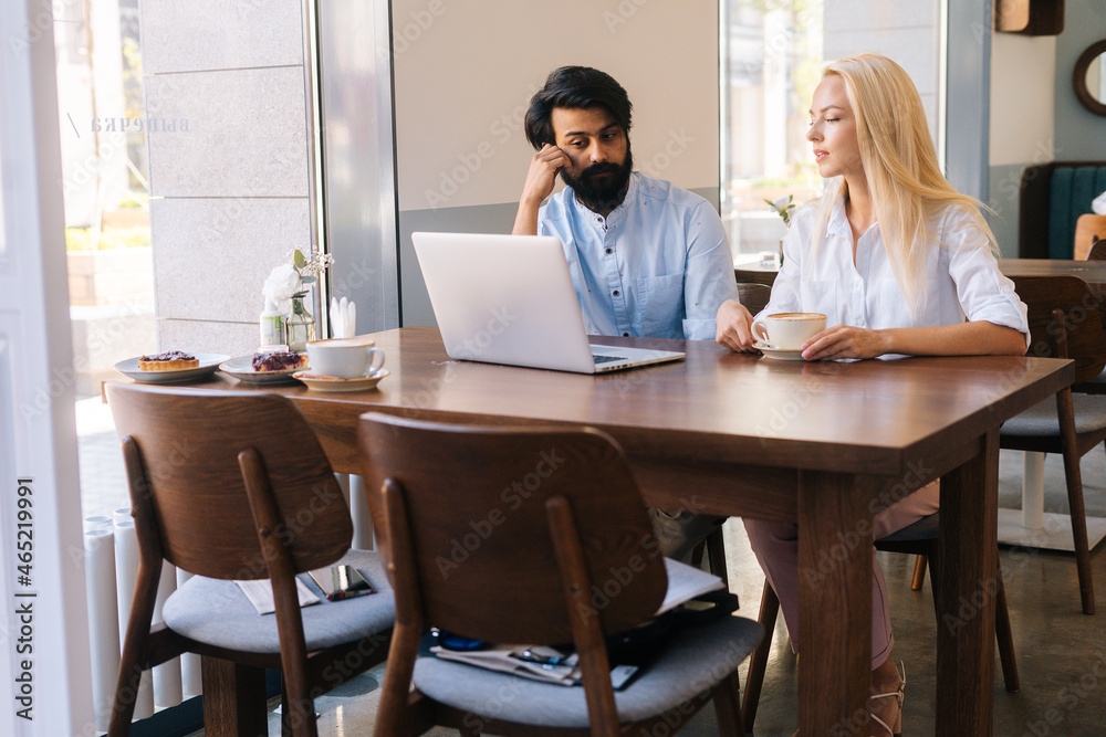 Portrait of young focused businesswoman and businessman working together using laptop sitting at cafe by window and drinking coffee. Two business partners or colleagues working in coffee shop.