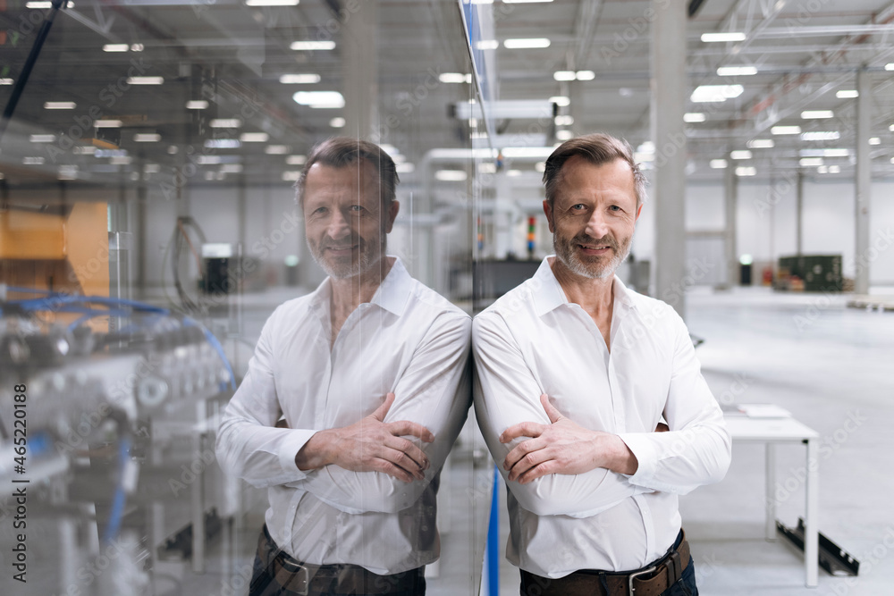 Smiling businessman standing with arms crossed while leaning on glass in factory Smiling businessman standing with arms crossed while leaning on glass in factory