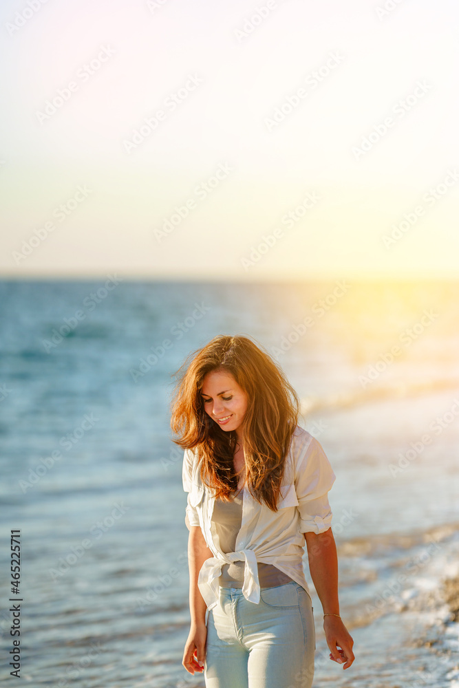 A charming young woman in a white shirt and jeans on a calm romantic beach by the sea. Summer woman vacations concept. The concept of active life. A charming young woman in a white shirt and jeans on a calm romantic beach by the sea. Summer woman vacations concept. The concept of active life.