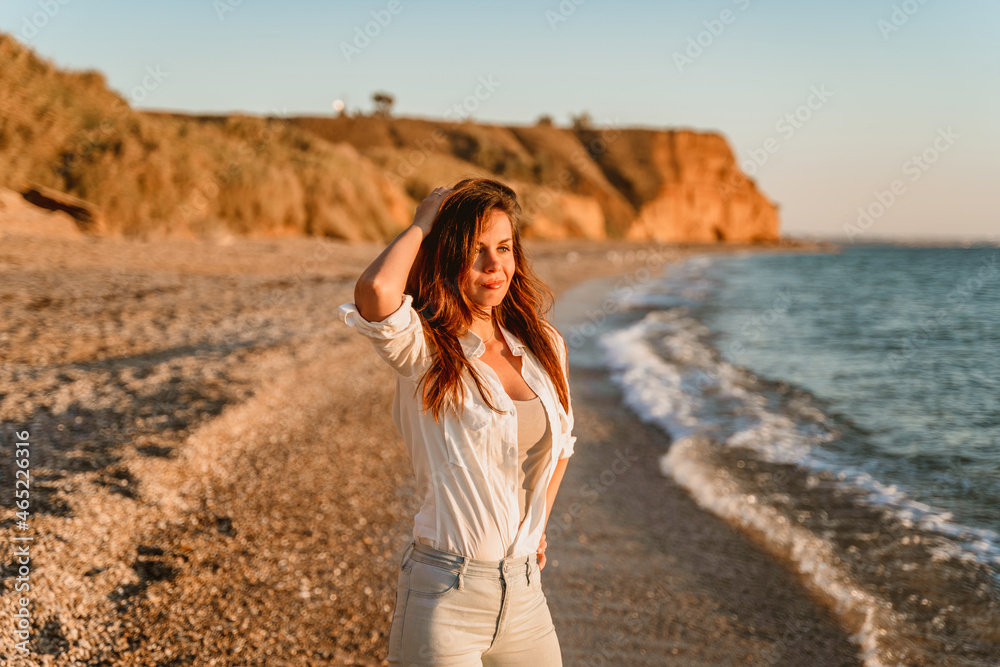 A charming young woman in a white shirt and jeans on a calm romantic beach by the sea. Summer woman vacations concept. The concept of active life. A charming young woman in a white shirt and jeans on a calm romantic beach by the sea. Summer woman vacations concept. The concept of active life.