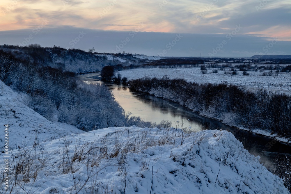 A beautiful winter view from the hill to the river with frosty trees growing along the banks. Winter landscape at sunset