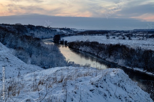 Fototapeta Naklejka Na Ścianę i Meble -  A beautiful winter view from the hill to the river with frosty trees growing along the banks. Winter landscape at sunset