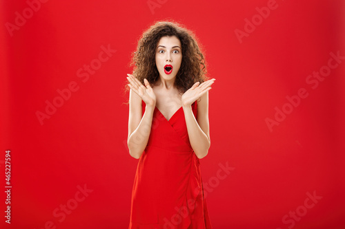 Woman learning surprising news being intrigued to hear all dirty ditails folding lips gasping making wow sound with raised palms in amazement gazing entertained and shocked at camera over red wall photo