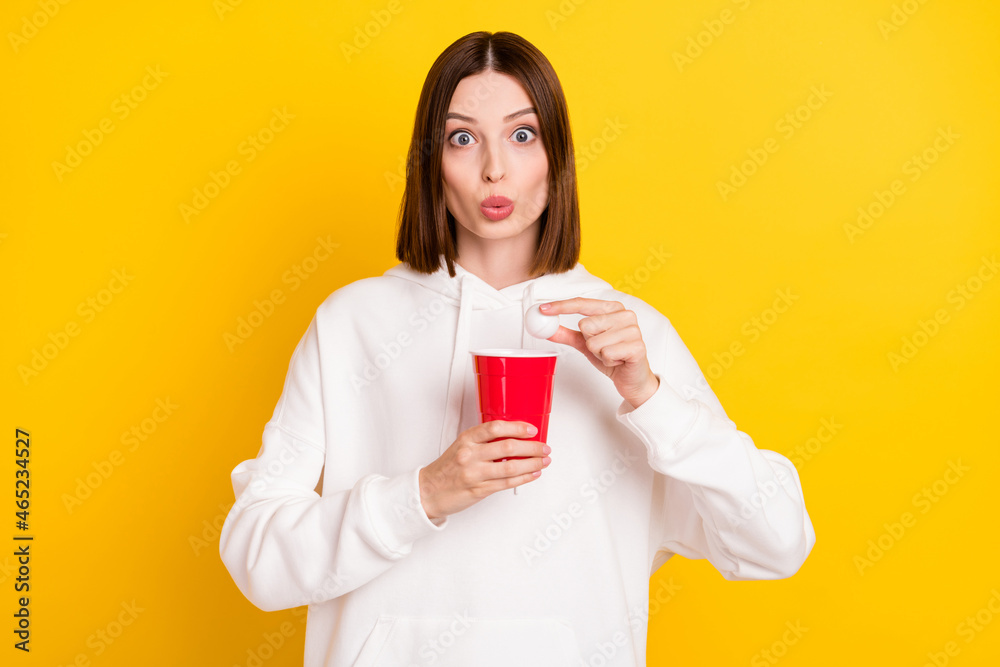 Photo of shocked lady hold alcohol beer cup ping pong ball wear white hoodie isolated yellow color background Photo of shocked lady hold alcohol beer cup ping pong ball wear white hoodie isolated yellow color background