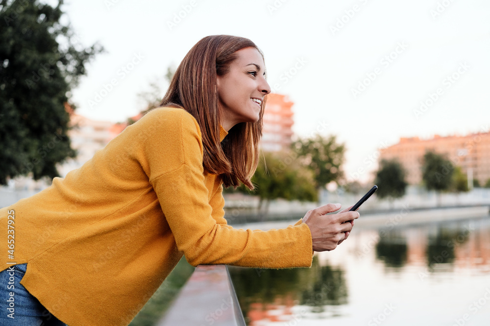 smiling caucasian woman wearing yellow pullover using mobile phone device outdoors in park during sunset. Technology and lifestyle smiling caucasian woman wearing yellow pullover using mobile phone device outdoors in park during sunset. Technology and lifestyle