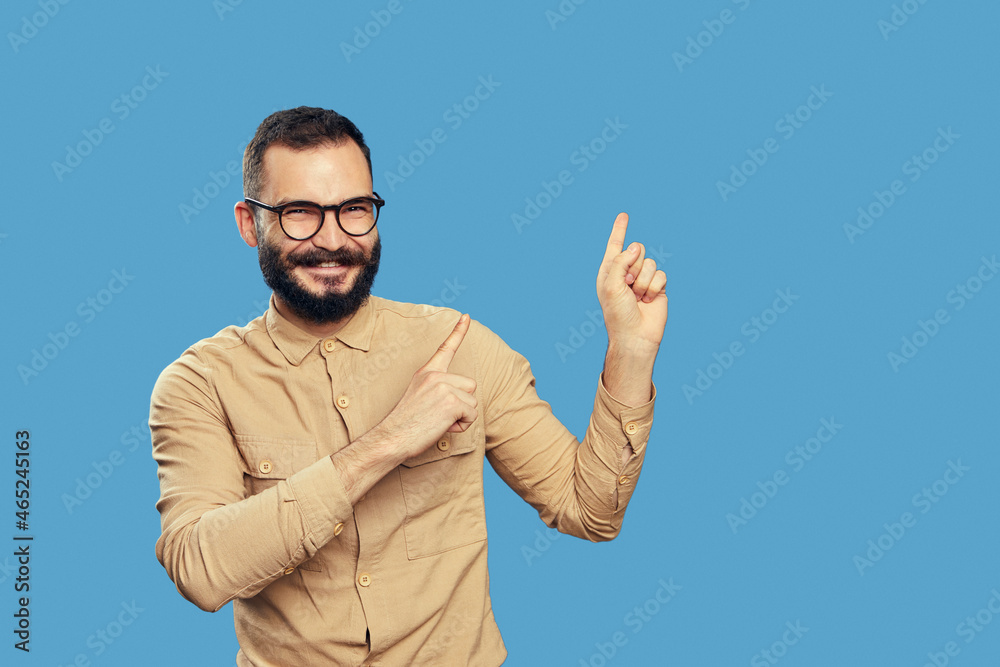 Naklejka premium Excited man points aside on empty space with both forefingers, advertises copy space for your advertising content, wears beige shirt, eyeglasses