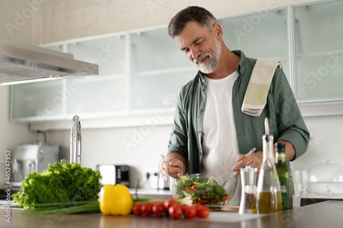 Happy mature man mixing a fresh vegetable salad standing in the kitchen at home.