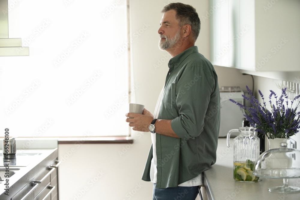 Happy mature man drinking coffee at home in the kitchen, enjoying hot drink in the morning on weekend.