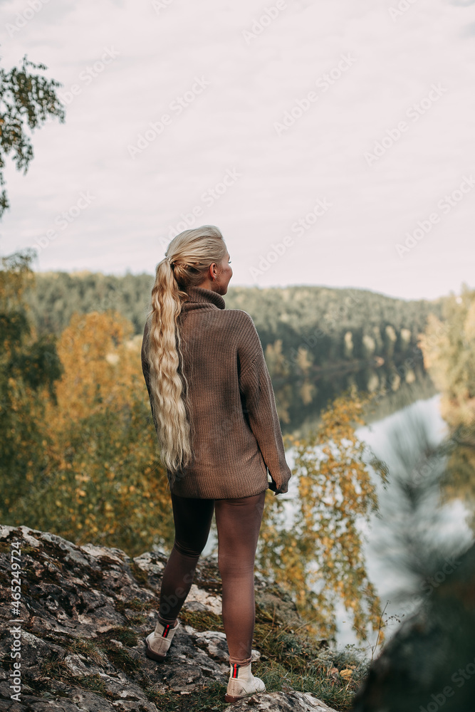 A young blonde girl in a brown hiking suit, in the autumn sometimes walks along the mountain with a view of the river and forest