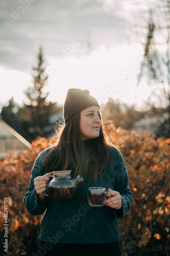 Portrait of a brunette girl with long hair in a dark green sweater and a gray hat outdoors in autumn with a glass teapot in her hands and a mug with tea