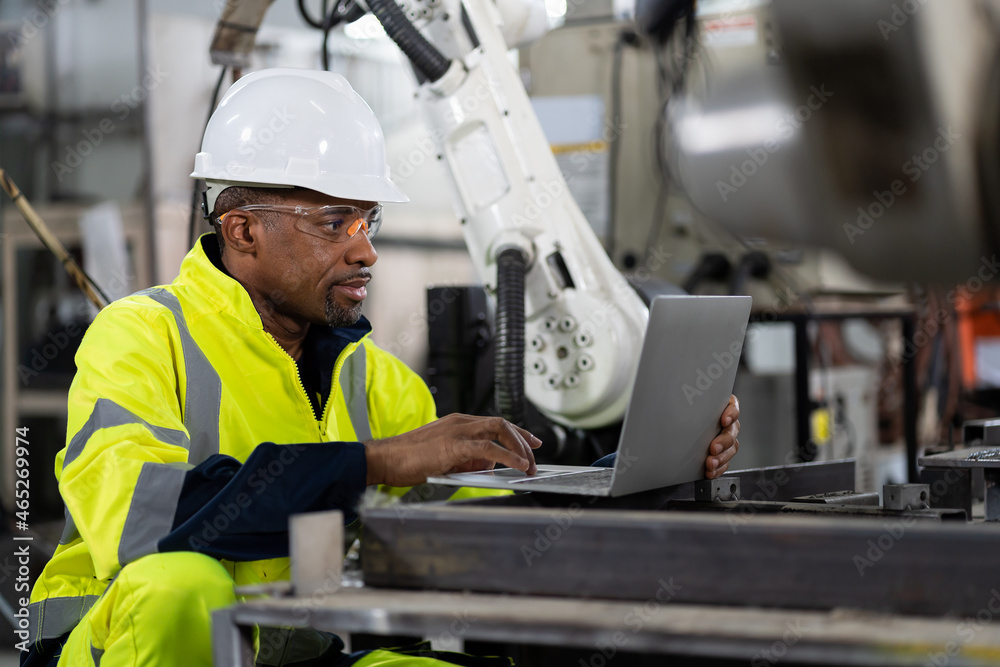 Male engineer worker using computer notebook control automatic robotic hand machine in factory. Black male technician worker working with control automatic robot arm system welding Male engineer worker using computer notebook control automatic robotic hand machine in factory. Black male technician worker working with control automatic robot arm system welding