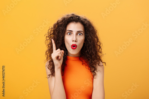 I got excellent plan. Enthusiastic emotive and excited female with curly hairstyle in orange cropped top raising index finger in eureka gesture folding lips popping eyes adding amazing suggestion photo