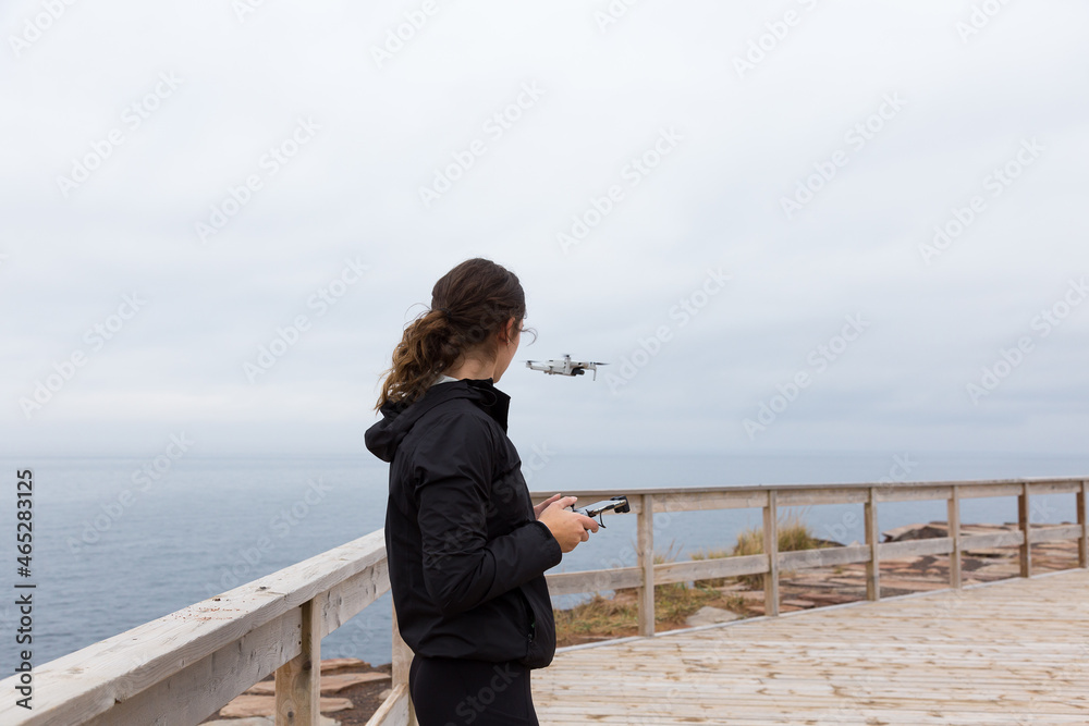 Medium back view of slim brunette young woman flying a drone near the coastline during a grey fall day, Cap-aux-Meules island, Magdalen Islands, Quebec, Canada