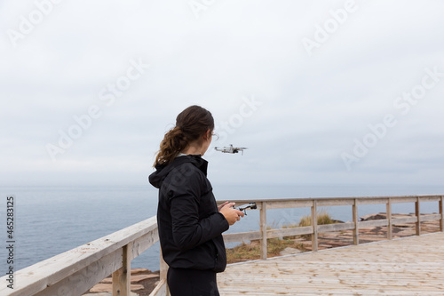 Medium back view of slim brunette young woman flying a drone near the coastline during a grey fall day, Cap-aux-Meules island, Magdalen Islands, Quebec, Canada