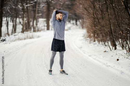 Fit sportsman doing stretching and warm up exercises while standing on snowy path in forest at winter. Healthy life, winter fitness, cold weather