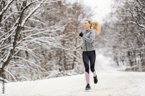 Full length of happy sportswoman jogging in nature on snowy path at winter. Healthy habits, outdoor fitness, winter fitness