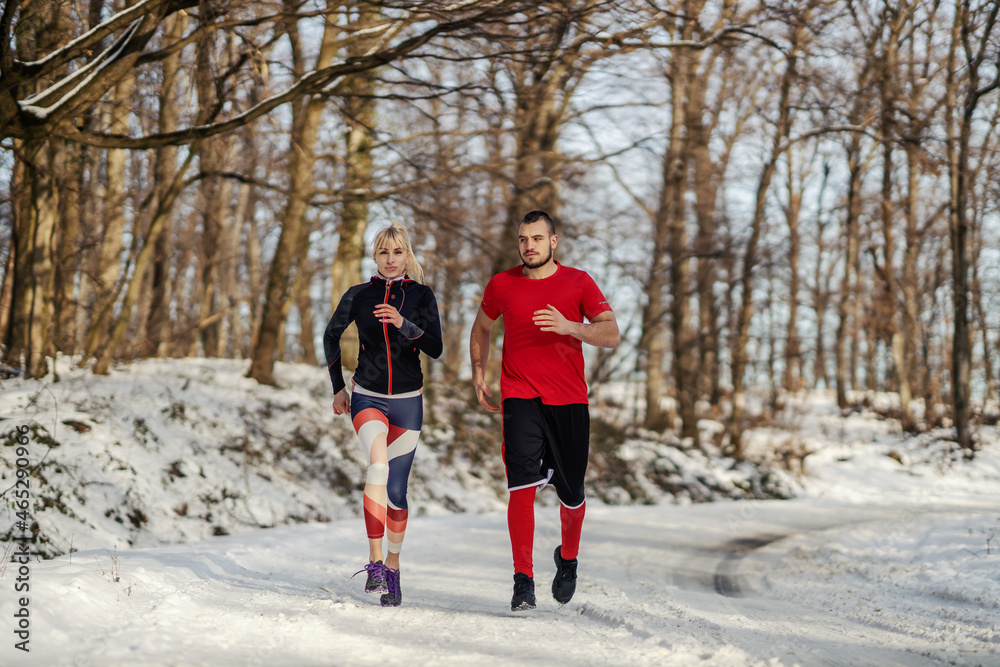 Naklejka premium Sporty couple jogging together in nature at snowy winter day. Relationship, winter fitness, cardio exercises