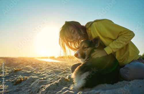 Happy woman in morning sun light with her pet at sandy beach. Girl hugging cute Welsh Corgi dog