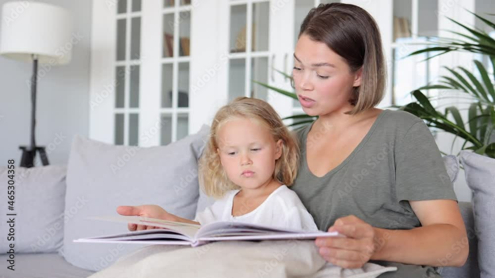 Mother and daughter education at home. spbi Young mum with stylish hairstyle reads fiction book to cute attentive little girl in living room close view Mother and daughter education at home. spbi Young mum with stylish hairstyle reads fiction book to cute attentive little girl in living room close view