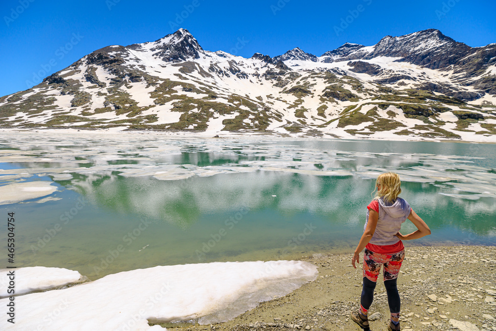 Woman trekking in the snow by reflecting White lake or Lago Bianco with icebergs in Switzerland. Lakefront on iced White lake in Grisons Canton at the Bernina pass by Sassal Mason mount.