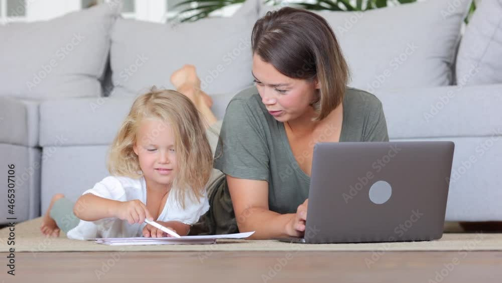 Mother with daughter at remote work from home. Cheerful young mommy freelancer uses spbi notebook computer while cute girl draws pictures on floor in living room Mother with daughter at remote work from home. Cheerful young mommy freelancer uses spbi notebook computer while cute girl draws pictures on floor in living room