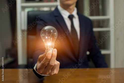 Businessman holding a bright light bulb saving bank a coins on wooden table 