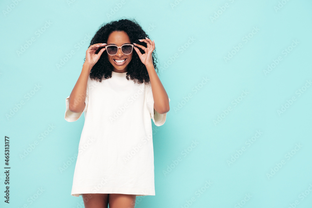 Beautiful black woman with afro hairstyle. Smiling model in long t-shirt clothes. Sexy carefree female posing near blue wall in studio. Tanned and cheerful in sunglasses Beautiful black woman with afro hairstyle. Smiling model in long t-shirt clothes. Sexy carefree female posing near blue wall in studio. Tanned and cheerful in sunglasses